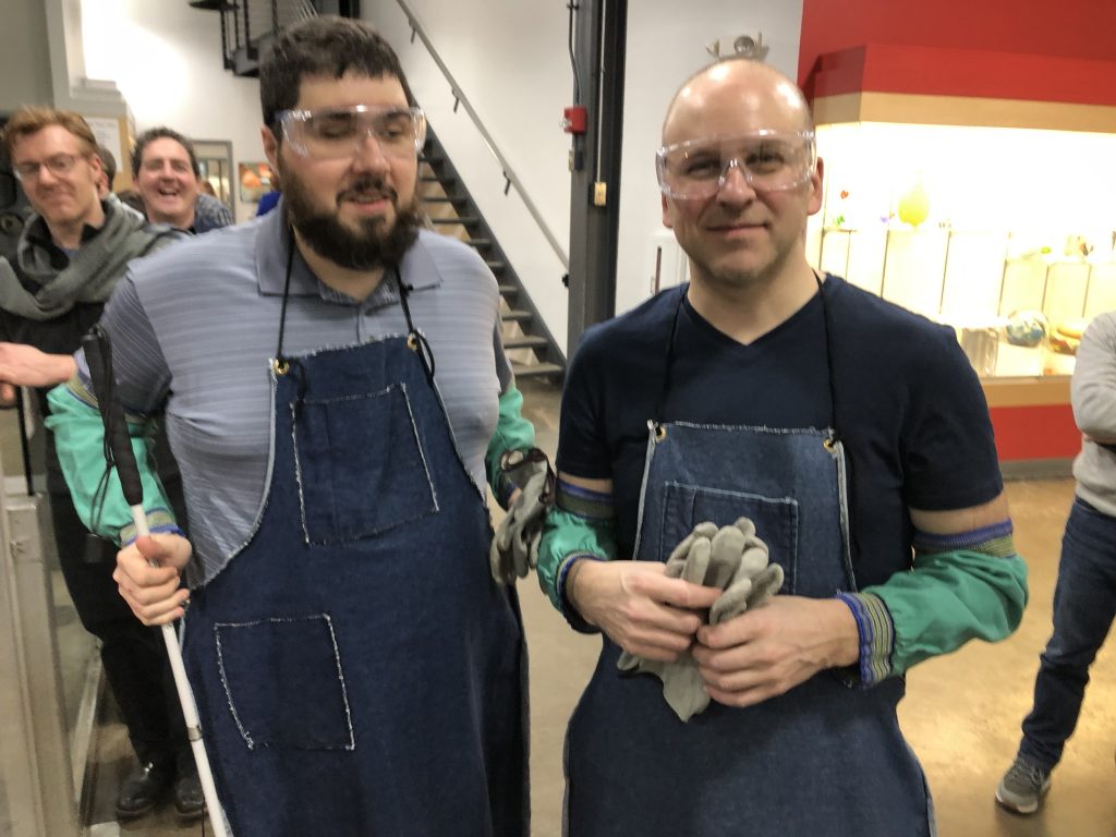 Sina, on left, and Corey, on the right, are both standing and facing the camera, wearing aprons, safety glasses, protective sleeves, and holding gloves.
