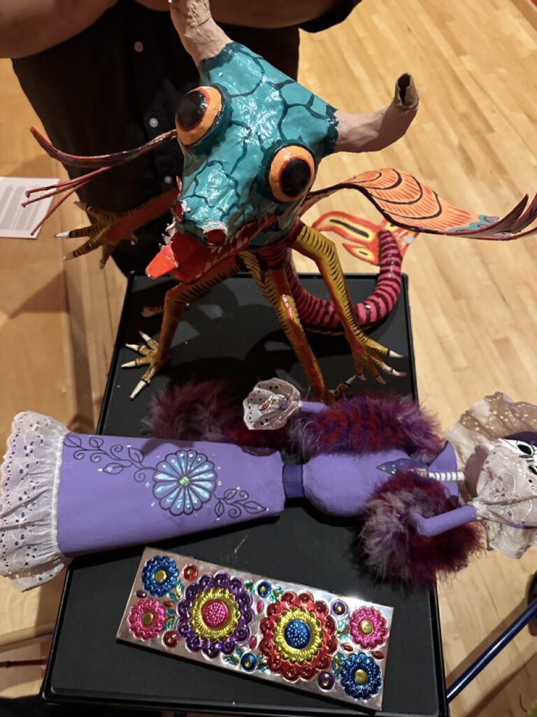 A colorful and eclectic collection of item. At the center, there is a whimsical creature with a turquoise textured body, large orange eyes with black pupils, and red accents around the eyes. It has a bright red mouth, long white tusks, and a pair of antlers. The creature's neck is adorned with yellow and red striped patterns, and it has multiple limbs in various colors, including yellow, red, and pink, with some resembling tentacles. To the left, there is a purple arm-like object with a hand at the end, featuring a lace cuff at the wrist and a floral pattern with a gem in the center on the upper part of the arm. Below, there is a rectangular object with a shiny, reflective surface, decorated with colorful gem-like structures in patterns of red, blue, green, and yellow, with silver accents. To the right, there is another arm-like object with a hand, similar to the one on the left, but this one is holding a white mask with a lace trim. The background is nondescript, and there is a partial view of a person wearing a black top. The items are placed on a black surface, possibly a table.